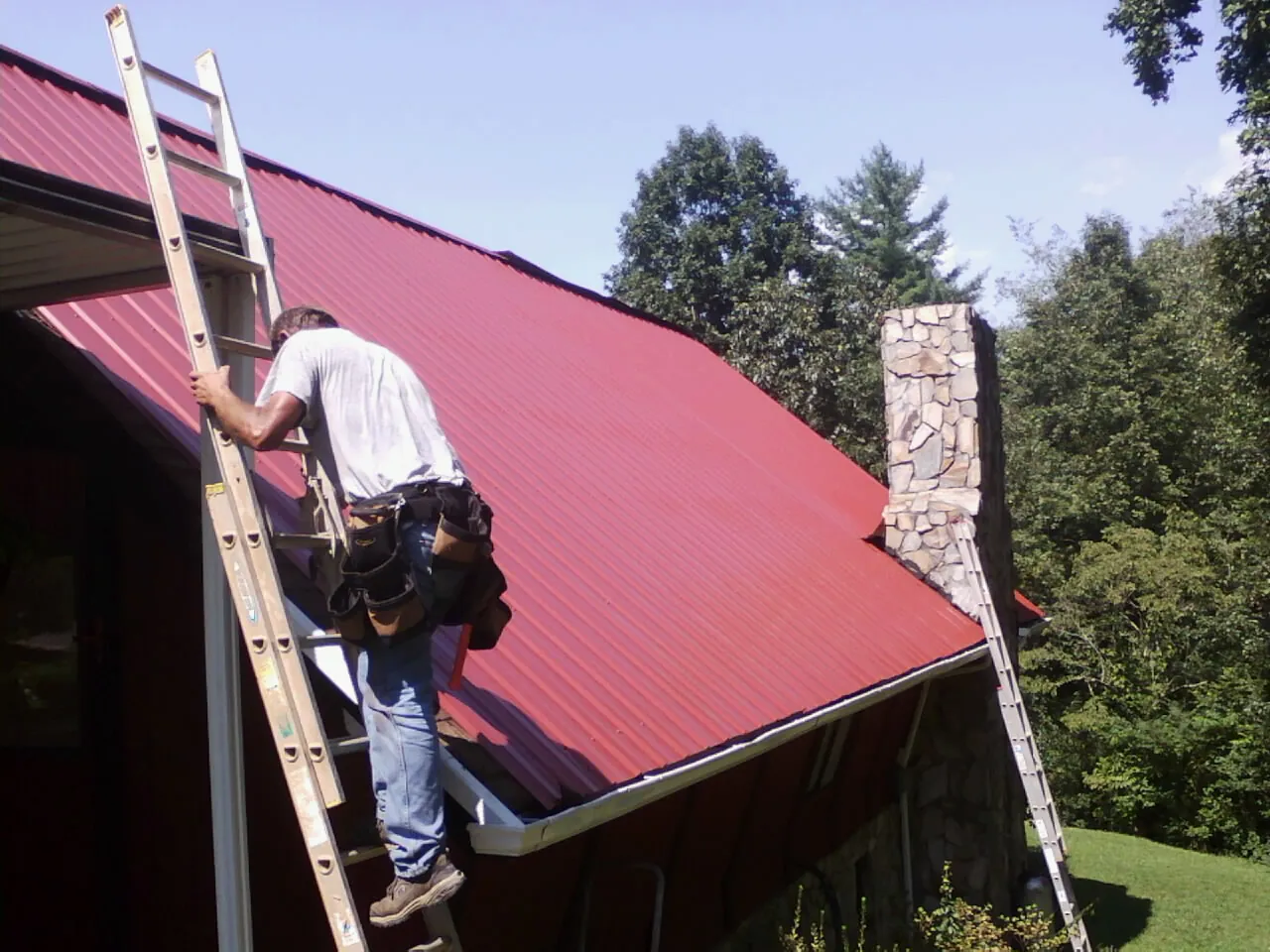Roofer on a ladder beside a red metal roof and stone chimney — Soffit & Fascia Repair by Roofing Brothers in West Decatur
